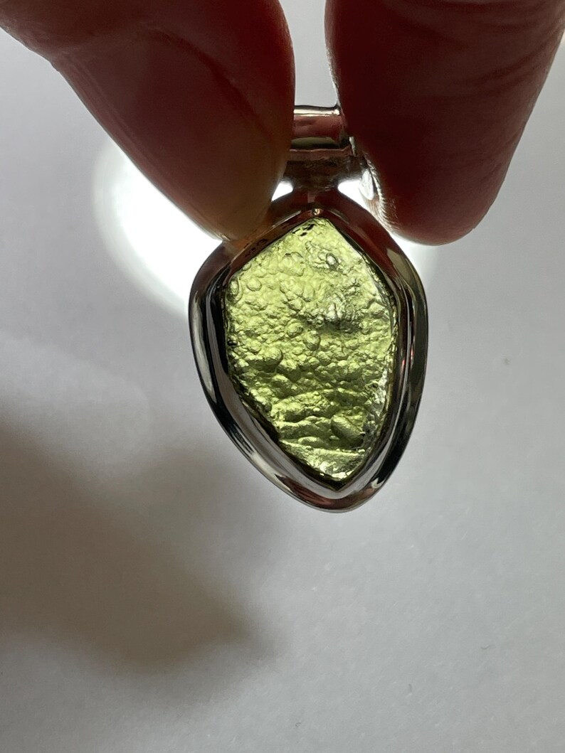 May include: A pendant featuring a raw, textured, olive-green moldavite stone set in a polished silver-toned metal frame. The stone has a pitted, irregular surface with a translucent quality, held within a smooth, teardrop-shaped metal bezel setting.