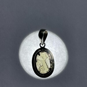 Puede incluir: Un colgante de plata con una piedra ovalada verde claro engastada en un bisel de plata. La piedra tiene un patr&oacute;n veteado &uacute;nico.