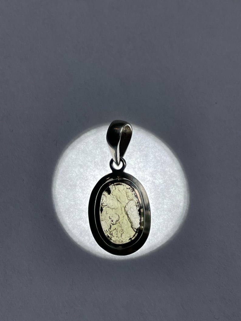 Puede incluir: Un colgante de plata con una piedra ovalada verde claro engastada en un bisel de plata. La piedra tiene un patr&oacute;n veteado &uacute;nico.