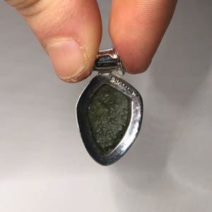 May include: A teardrop-shaped pendant featuring a textured, olive-green stone set in a polished silver-toned metal frame. The metal bail at the top is stamped with the text 925, indicating the metal composition of the setting for this jewelry piece.