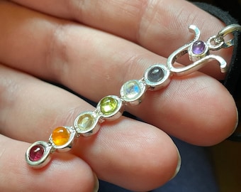 May include: A silver necklace with seven round gemstones in the colors of the rainbow. The stones are arranged in a line and are separated by small silver beads. The necklace has a silver clasp at the top.