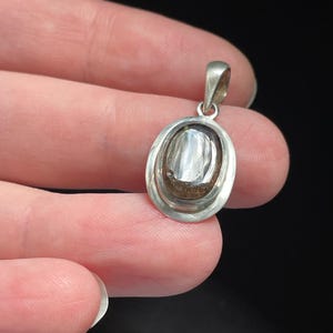 Puede incluir: Un colgante ovalado de plata con una piedra transparente en su interior. La piedra tiene un color gris oscuro y una superficie texturizada.