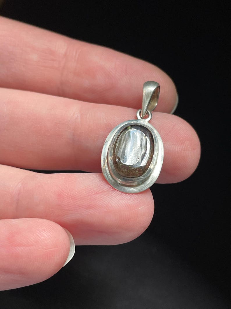 Puede incluir: Un colgante ovalado de plata con una piedra transparente en su interior. La piedra tiene un color gris oscuro y una superficie texturizada.