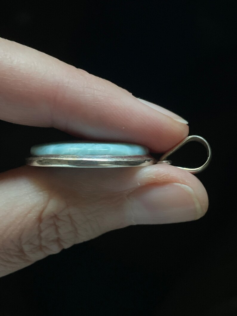 Puede incluir: Un colgante de plata con una piedra preciosa azul claro engastada en un bisel de plata. El colgante tiene una simple anilla para sujetarlo a una cadena.