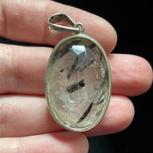 May include: Silver pendant featuring a clear quartz stone with black tourmaline inclusions. The pendant is oval shaped and has a polished finish.