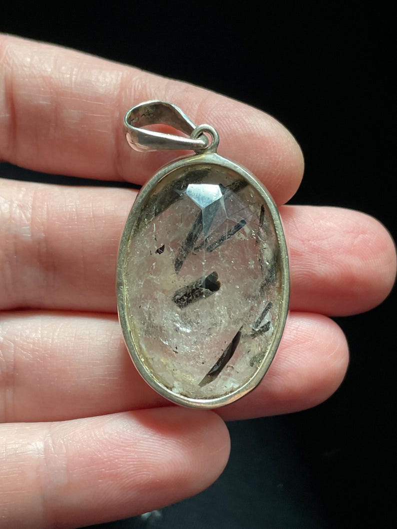 May include: Silver pendant featuring a clear quartz stone with black tourmaline inclusions. The pendant is oval shaped and has a polished finish.