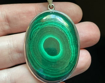 May include: A silver pendant with a large oval-shaped green malachite stone. The stone has a swirling pattern of concentric circles.