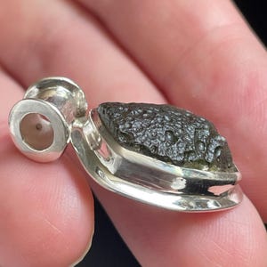May include: A pendant featuring a rough, dark olive-green moldavite stone with a textured, pitted surface. The stone is securely set in a polished silver bezel with a smooth, cylindrical silver bail attached to the top for a necklace chain.