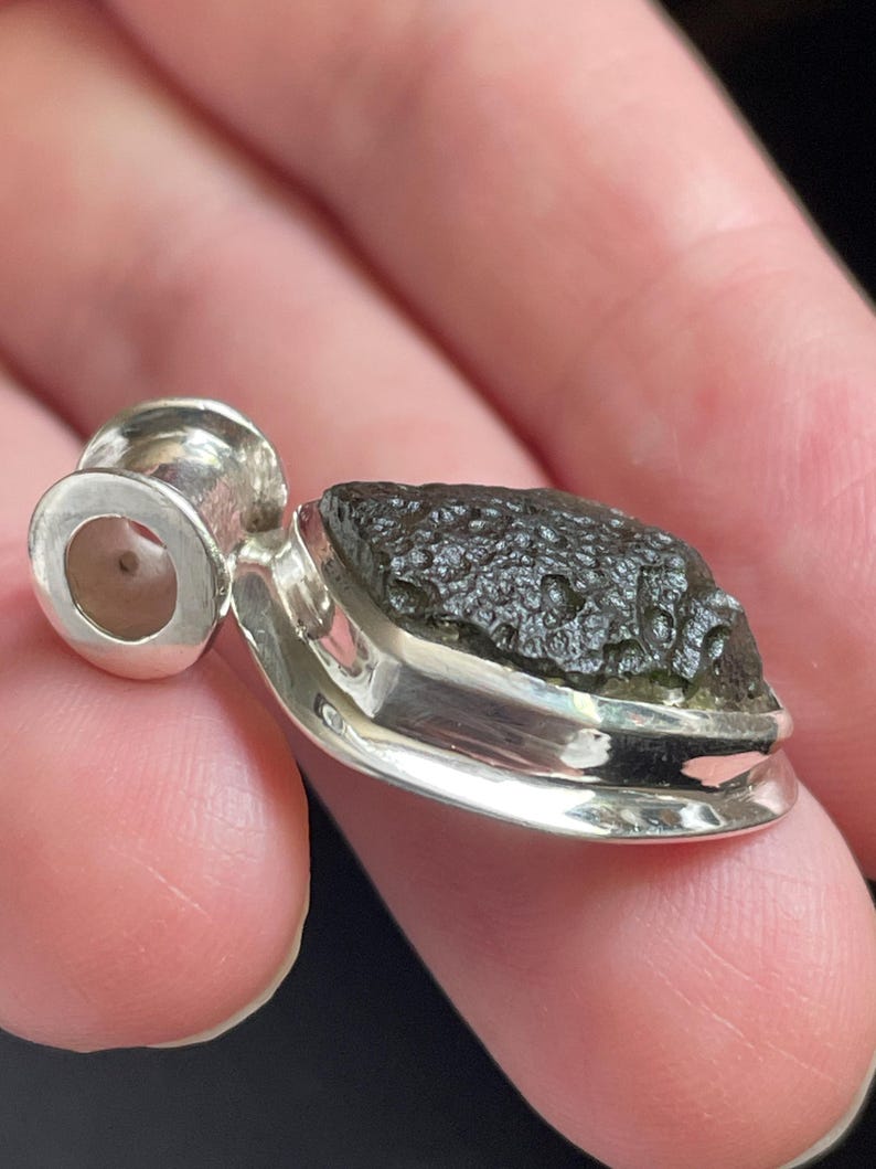 May include: A pendant featuring a rough, dark olive-green moldavite stone with a textured, pitted surface. The stone is securely set in a polished silver bezel with a smooth, cylindrical silver bail attached to the top for a necklace chain.