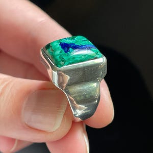 K&ouml;nnte beinhalten: Ein Silberring mit einem rechteckigen, polierten Azurit-Malachit-Edelstein. Der Stein zeigt lebendige Wirbel in tiefem Blau und hellem Gr&uuml;n. Das Metallband ist glatt, dick und hat eine hochgl&auml;nzende, reflektierende Oberfl&auml;che um den Stein herum.