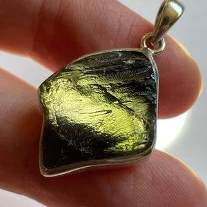 May include: A silver pendant featuring a large, irregularly shaped green and black gemstone. The gemstone has a rough, natural texture and is set in a silver frame.
