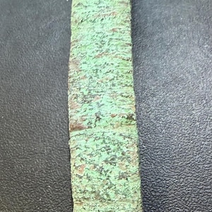 May include: A rectangular metal bar with a weathered, oxidized surface featuring a prominent mint green patina. The object has a rough, textured appearance with visible brown and metallic undertones throughout its elongated, narrow, and flat form.