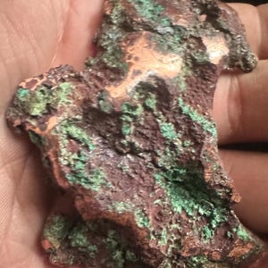 May include: A close-up of a copper ore specimen with a rough, irregular shape. The surface displays a mix of reddish-brown, green, and copper-colored patches, showcasing natural oxidation and mineral formations. The texture appears coarse and uneven, highlighting the raw, unrefined nature of the material.