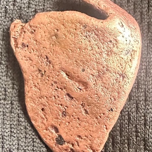 May include: A reddish-brown, heart-shaped object with a textured surface and a curved handle. The object appears to be made of a solid material, possibly metal or stone. The shape is organic and irregular.