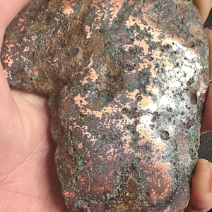May include: A rough, irregularly shaped copper ore specimen with a mix of brown, green, and metallic copper hues. The surface shows a textured, weathered appearance, with areas of oxidation and varying shades of color. The ore is held in a hand.