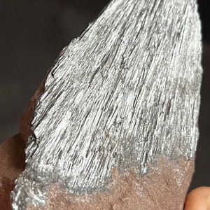 May include: A close-up of a mineral specimen featuring a silver-colored, fibrous structure atop a reddish-brown base. The metallic fibers have a striated appearance, creating a textured surface. The rock is irregularly shaped.