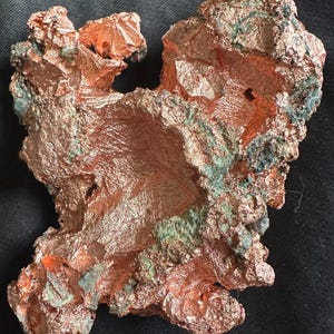 May include: A raw copper specimen with a rough, textured surface. The copper has a reddish-brown color with patches of green and blue-green oxidation. The mineral formation has an irregular shape with multiple protrusions and cavities.