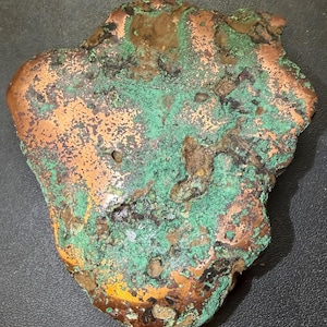 May include: A raw, irregular piece of copper metal featuring a natural, oxidized patina with vibrant green and turquoise mineral deposits contrasting against the smooth, metallic copper-colored surface and dark, textured recessed areas.