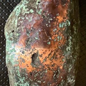 May include: An irregular, elongated object with a metallic copper surface showing significant oxidation. The piece features a mix of smooth, reflective reddish-brown copper areas and rough, textured patches of light green and gray patina.