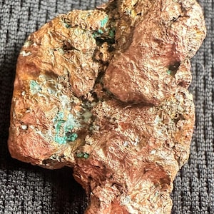 May include: A raw copper specimen with a rough, textured surface. The copper has a reddish-brown color with patches of green and white minerals. The overall shape is irregular and organic, suggesting a natural formation.