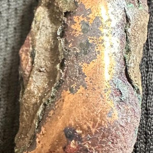 May include: A weathered, irregularly shaped object with a copper-colored surface. Patches of green and dark brown hues are visible, suggesting oxidation. The object has a rough, textured appearance.