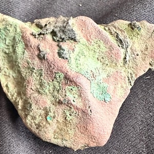 May include: A weathered, irregularly shaped stone fragment with a mix of colors. The surface displays patches of pink, green, and tan hues, with a rough, textured appearance. The fragment has a jagged edge and appears to be an ancient artifact.