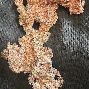 May include: A raw, irregularly shaped copper specimen with a textured, porous surface. The copper has a reddish-brown color with hints of gold and green. The piece has several holes and a rough, organic appearance, suggesting it is a natural mineral formation.