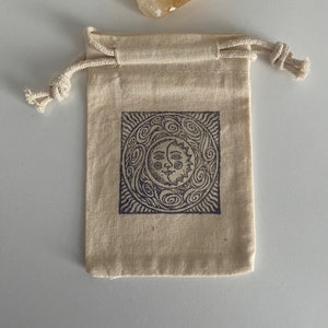 May include: A small, beige drawstring bag with a sun and moon design printed on the front. The bag is made of natural fabric and has a simple, minimalist style.