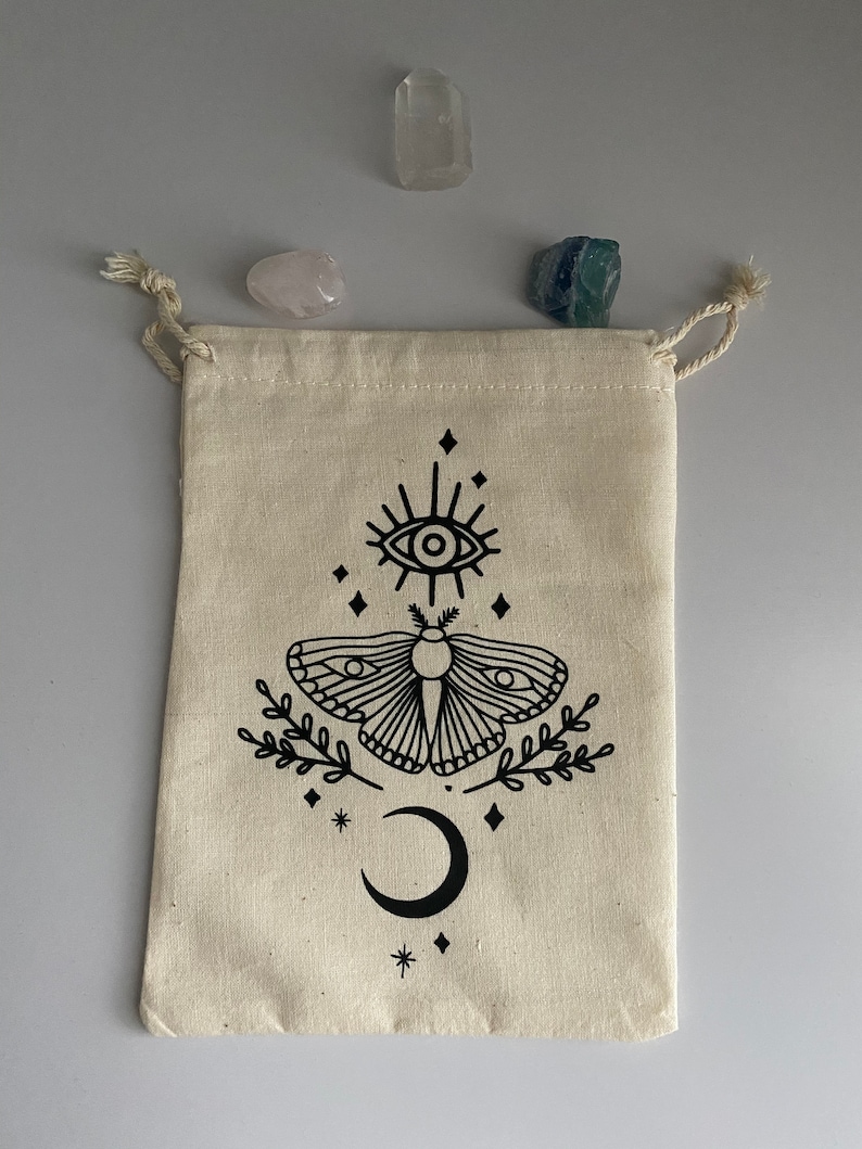 May include: A white cotton drawstring bag with a black and white illustration of a moth, a crescent moon, and a star. The bag is decorated with a black eye with a starburst design.