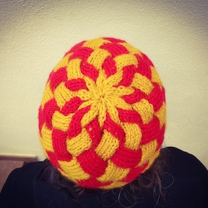 May include: A red and yellow knitted hat with a circular pattern. The hat is made of yarn and has a textured surface.