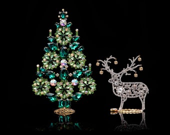 May include: A jeweled Christmas tree and reindeer figurine. The tree is made of green and clear crystals, with a gold-colored base. The reindeer is silver-colored with gold-colored accents. Both are set against a black background.