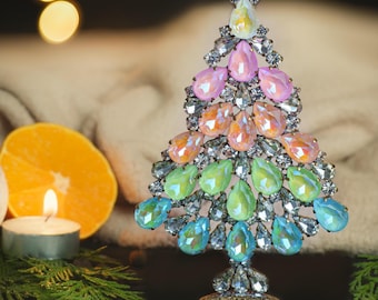 May include: A decorative Christmas tree ornament with a silver metal frame and colorful, teardrop-shaped gemstones. The gemstones are arranged in a gradient of colors, including blue, green, orange, and pink. The tree is approximately 6 inches tall.