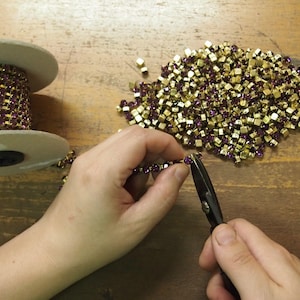 May include: A person is using a pair of pliers to cut a chain of gold and purple square-shaped rhinestones. The chain is on a spool and there is a pile of loose rhinestones on the table.