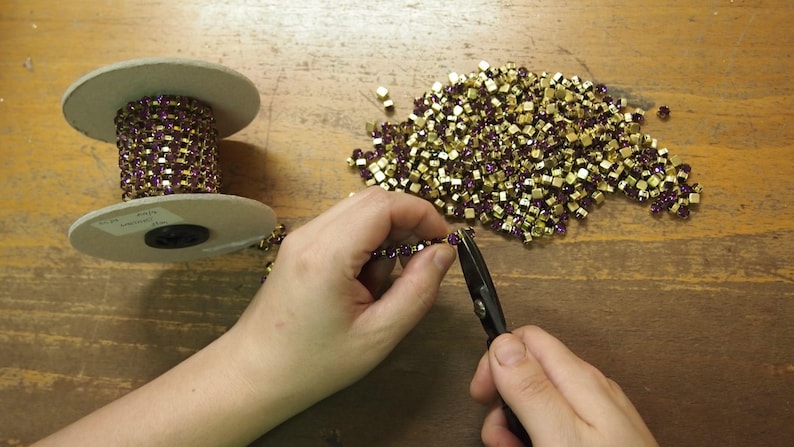 May include: A person is using a pair of pliers to cut a chain of gold and purple square-shaped rhinestones. The chain is on a spool and there is a pile of loose rhinestones on the table.