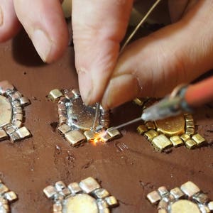May include: A close-up of a person soldering small, intricate metal pieces together on a brown clay surface. The pieces are arranged in a pattern and appear to be part of a larger design.