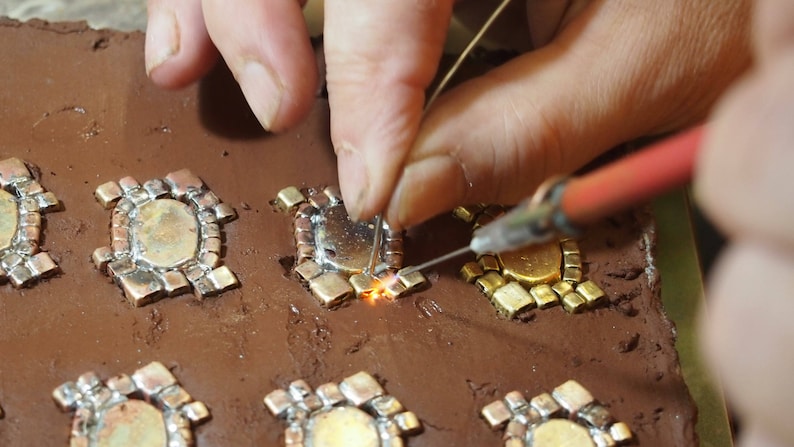 May include: A close-up of a person soldering small, intricate metal pieces together on a brown clay surface. The pieces are arranged in a pattern and appear to be part of a larger design.