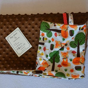 May include: A brown and white baby blanket with a fox print. One side of the blanket is brown with a textured surface and the other side is white with a fox print. The blanket has a tag that says "Baby Blanket".