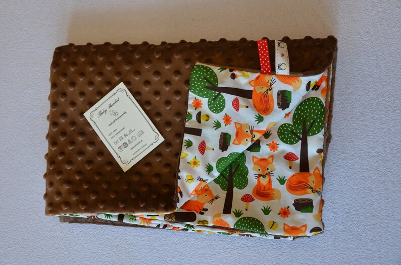 May include: A brown and white baby blanket with a fox print. One side of the blanket is brown with a textured surface and the other side is white with a fox print. The blanket has a tag that says "Baby Blanket".