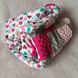 May include: A folded baby blanket featuring a heart pattern in pink, red, gray, and teal on a white background. The blanket has a pink minky dot fabric and gray minky dot fabric. The blanket is a patchwork design.