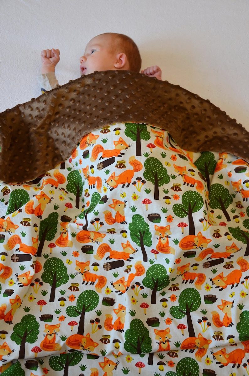 May include: A brown and white baby blanket with a fox print. The blanket is made of soft, plush fabric and features a brown minky backing. A baby is lying on the blanket.