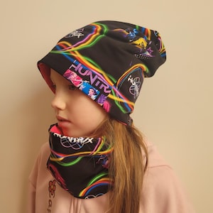 Könnte beinhalten: Schwarzes Mütze- und Halstuch-Set mit Neon-Design. Die Mütze trägt das Wort "HUNTR" und eine farbenfrohe Grafik. Das Halstuch spiegelt das Design wider und bildet ein passendes Set. Das Set ist für kaltes Wetter konzipiert.