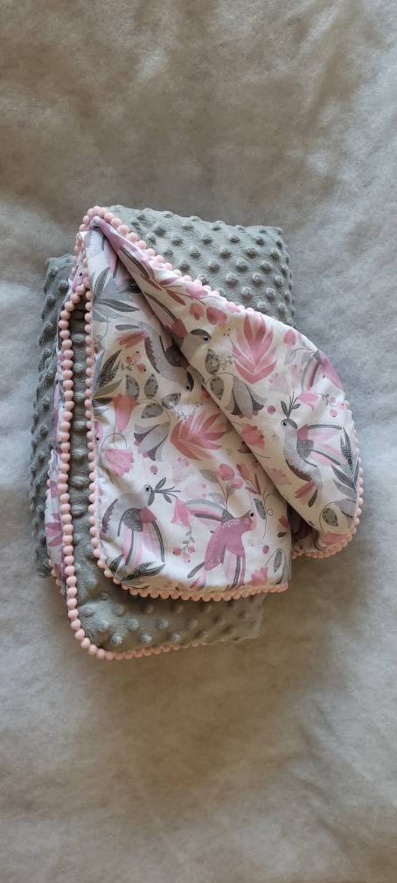 Pode incluir: Um cobertor de beb&eacute; cinzento e rosa com um padr&atilde;o floral e uma guarni&ccedil;&atilde;o de pompons brancos. O cobertor &eacute; feito de tecido macio e felpudo.