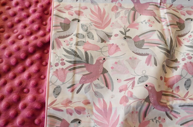 Pode incluir: Um cobertor para beb&eacute; rosa e branco com um padr&atilde;o de p&aacute;ssaros e flores. O cobertor tem uma textura macia e felpuda de um lado e uma textura lisa de algod&atilde;o do outro lado.