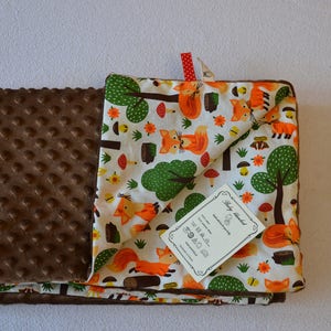 May include: A brown and white baby blanket with a fox and forest print on one side and a brown minky dot fabric on the other side. The blanket has a tag that says "Baby Blanket".