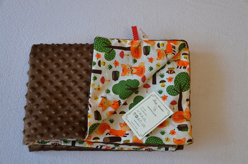 May include: A brown and white baby blanket with a fox and forest print on one side and a brown minky dot fabric on the other side. The blanket has a tag that says "Baby Blanket".