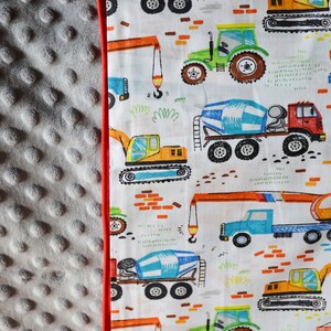 May include: A white fabric with a red border features a repeating pattern of construction vehicles. The vehicles are drawn in a cartoon style and include cement mixers, tractors, excavators, and cranes. The background is white with orange brick patterns.