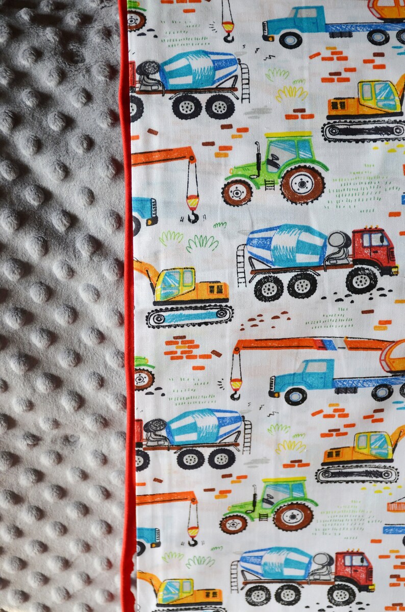 May include: A white fabric with a red border features a repeating pattern of construction vehicles. The vehicles are drawn in a cartoon style and include cement mixers, tractors, excavators, and cranes. The background is white with orange brick patterns.