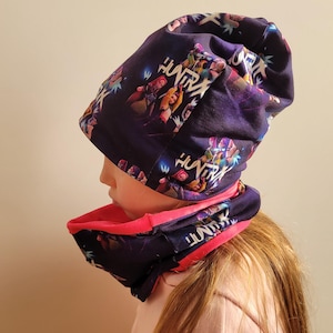May include: A dark purple beanie and neck gaiter set with a vibrant print featuring characters and the word "HUNTRA". The interior of the neck gaiter is bright pink. The beanie is worn on a person.