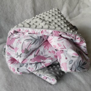 Pode incluir: Um cobertor de beb&eacute; cinzento e rosa com uma superf&iacute;cie macia e texturizada e um padr&atilde;o floral com p&aacute;ssaros cor-de-rosa. O cobertor est&aacute; dobrado ao meio e tem uma borda branca.