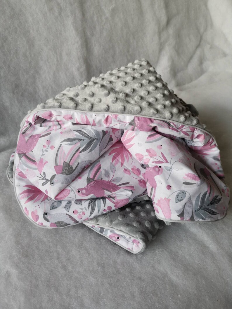 Pode incluir: Um cobertor de beb&eacute; cinzento e rosa com uma superf&iacute;cie macia e texturizada e um padr&atilde;o floral com p&aacute;ssaros cor-de-rosa. O cobertor est&aacute; dobrado ao meio e tem uma borda branca.